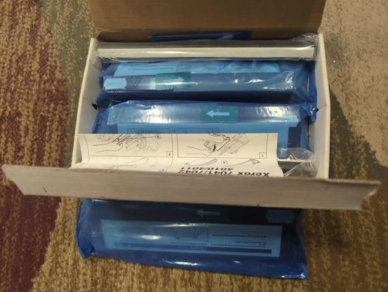 The same box and blue plastic bag, but the box is now open, showing two more blue plastic bagged oblong items inside it, as well as a folded piece of paper with diagrams of hands installing toner cartridges. Verox 7041/7042/4010/4011 is printed at the top of the page.