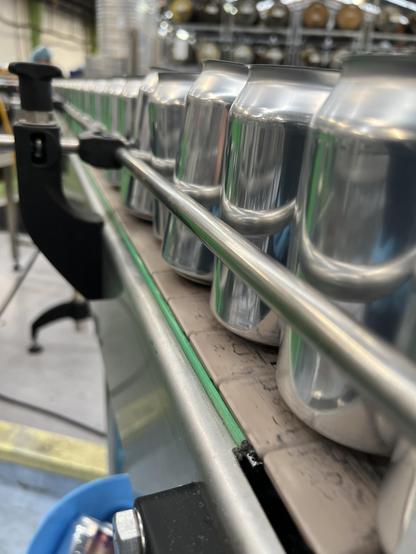 A dramatic perspective shot of a long row of 330mL beverage cans on a conveyor track with rails. Various brewery related stuff is in soft focus in the background.