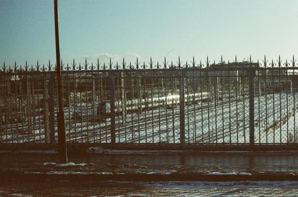 A film photography taken from a bridge over some train tracks covered in snow. The guardrail is visible in the foregroud, and a train is passing on the rails behind it. Sunlight is gleaming on the train and shining through the guardrail. The picture is grainy and slightly yellowish.