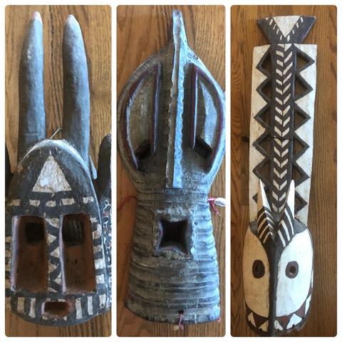 Three traditional African masks arranged side by side on a wooden surface. The left mask features elongated horns and tribal patterns. The center mask has a rounded shape with a star-like opening. The right mask shows a stylized face with geometric designs and prominent features.