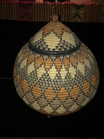 A decorative woven Zulu beer basket with a conical lid, featuring intricate geometric patterns in shades of brown, black, and tan. The background includes a colorful woven textile.