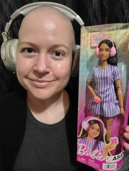 Woman in mid-30s with shaved head wearing gold over-ear headphones smiling happily and holding up an "Autistic Barbie" doll in its box.