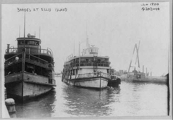 The image is a black-and-white photograph dated June 1920, titled "Barges at Ellis Island." It shows two large boats docked in a harbor. The boat on the left is named "Thomas C. Millase" and appears to be a tugboat or ferry, with a cabin structure and a few visible people on deck. The boat on the right is a larger passenger or ferry boat, with multiple decks and a higher cabin structure, suggesting it is designed to carry more passengers. There are several people visible on the upper deck of the larger boat, and it seems to be docked at a pier with a crane in the background, indicating industrial or port activity. The water reflects the boats and the surrounding structures, and the overall scene conveys a busy harbor environment typical of the early 20th century.