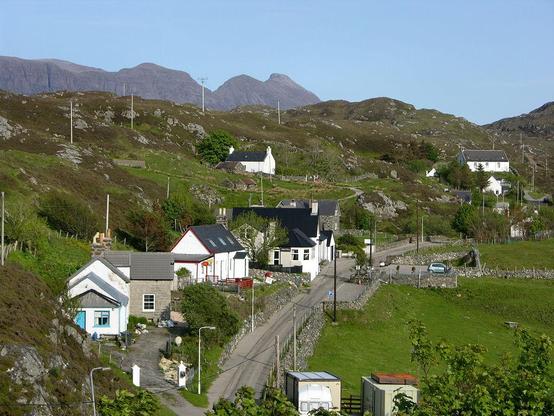 The village of Drumbeg. The image shows a view from above of a road leading away from the photographer past a scatter of buildings with lower ground to the right and high ground rising to the left behind the buildings. There's part of a larger mountain visible above the main skyline on the left. The scene is in sunshine.