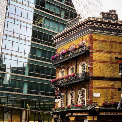 The Albert Pub: A charming, multi-story Victorian pub named "The Albert" built of yellow brick with red accents. It features black wrought-iron balconies overflowing with pink flowers, standing in stark contrast to the massive, modern glass skyscraper directly behind it.