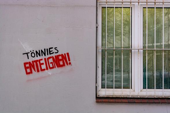 Links neben einem vergitterten Fenster steht an einer hellgrauen Häuserwand Tönnies Enteignen!