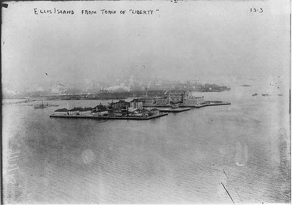 This image is a historical photograph of Ellis Island, as seen from the top of the Statue of Liberty. The photograph is in black and white, giving it an aged appearance. The island is surrounded by water, and the island itself has several buildings with a symmetrical layout. The architecture of the buildings suggests a utilitarian style, typical of the time period. In the background, there are smokestacks emitting smoke, indicating industrial activity, likely related to the processing of immigrants at Ellis Island. The sky appears overcast, adding to the somber tone of the image. The photograph is labeled at the top with the text "ELLIS ISLAND FROM TOP OF 'LIBERTY'" and has a number "13-3" in the top right corner, suggesting it may be part of a series or collection.