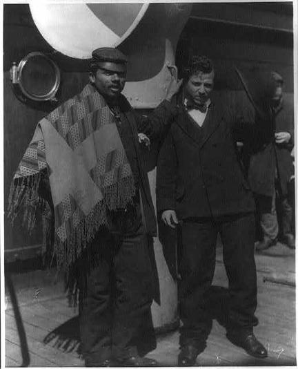 This image is a black-and-white photograph featuring two men standing on a ship. The man on the left is dressed in a traditional outfit that includes a patterned shawl or blanket draped over his shoulders and a cap. He appears to be of African descent and is standing with his hands at his sides. The man on the right is dressed in formal attire, wearing a suit with a vest, a tie, and a bowler hat. He is standing with his left arm resting on the shoulder of the man on the left, and his right hand is in his pocket. In the background, there is a large ship's wheel and a porthole, indicating that the photo was taken on a ship. The overall setting and attire suggest a historical context, possibly from the early 20th century.
