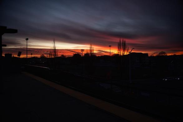 A colorful sunset with shades of orange, pink, and purple fills the sky, silhouetting trees and buildings. The scene captures a train platform in the foreground, enhancing the tranquil atmosphere.