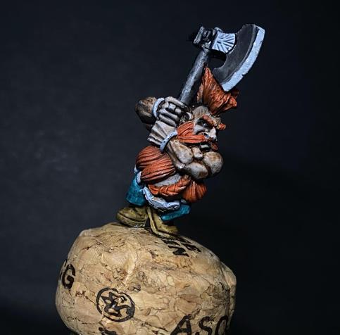 A dwarf slayer miniature stuck on a cork with some Contrast paints applied. Big bright orange crest and beard as it should be. Two hands on an axe.