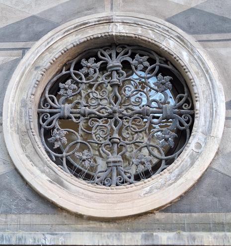 Links vom Eingang zum Museum ist ein ca 80 cm großes, rundes Fenster in die Wand eingelassen. Es hat einen breiten steinernen Rahmen mit barocken Elementen. Doch der Blickfang ist das reich verzierte, schmiedeeiserne Gitter vor dem Fenster. Es ist viergeteilt und über und über mit Blumen, Blättern und rund gebogenen Stängeln geschmückt.
