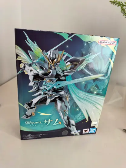 The image shows a product box for a Bandai Namco SHFiguarts figure. The box features artwork of a silver and green armored robot figure named "SAM" (with the Japanese text "サム" displayed), holding weapons and adorned with crystalline wings and flowing green accents. Text on the box includes "BANDAI NAMCO" in the top right corner, "SHFiguarts" and "SAM" (with "SAM" in English and "サム" in Japanese) near the bottom left, and "BANDAI" branding at the bottom right. The background of the artwork is dark blue with light effects, and the figure has detailed armor with gold and black accents. The box is positioned on a light-colored surface with a small plant visible in the top right corner.