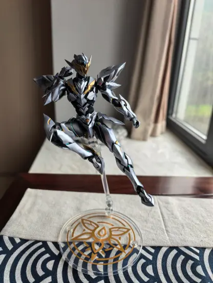 A detailed silver, gold, black, and blue armored action figure in a dynamic pose, mounted on a clear stand with a circular base featuring a gold floral design. The figure has sharp, angular armor segments and is positioned on a surface covered by a blue-and-white patterned fabric and a light gray cloth. Behind the figure, a window with brown curtains reveals an outdoor view of greenery, with a wall and dark wooden trim visible. The stand is transparent, and the base displays a decorative gold pattern with no text. The scene is set in a well-lit indoor space with natural light from the window.