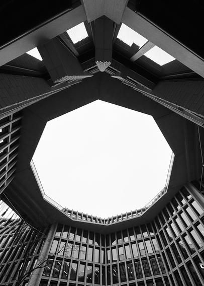 A black-and-white photograph showcasing the interior of a building with an octagonal opening at the top. The view includes angular architectural elements and large windows surrounding the space, creating a modern and unique design. The sky is visible through the opening, contributing to
