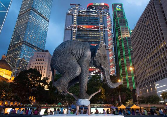 A sculpture of a large elephant standing on the back of a human figure, in a busy urban setting, surrounded by modern skyscrapers at dusk.