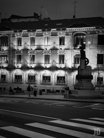 Fotografía en blanco y negro de una escena urbana nocturna. En el centro, un edificio de estilo clásico y ornamentado, iluminado dramáticamente desde abajo, lo que resalta sus detalles arquitectónicos y balcones de hierro forjado contra un cielo oscuro. La planta baja del edificio presenta varias ventanas arqueadas, algunas con letreros como "Seguros" y "Ocaso".

A la derecha del edificio, se alza una estatua oscura de una figura humana sobre un gran pedestal de piedra, que incluye inscripciones como "EMPRESARIO". En primer plano, una calle oscura con un paso de cebra visible. Varias personas caminan por la acera, incluyendo dos mujeres en el centro de la imagen y una persona de pie cerca de la estatua a la derecha. Algunos árboles desnudos se ven a lo largo de la acera.