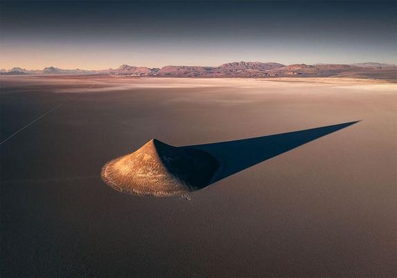 Aerial view of a conical volcanic formation casting a long shadow over a vast, flat landscape under a clear sky.