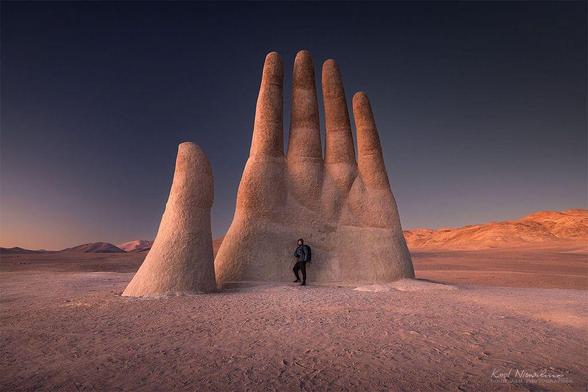 A giant hand sculpture rises from the desert landscape, with a person standing beside it against a twilight sky.