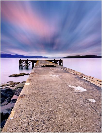 A long concrete pier extending into calm waters, with a smooth concrete surface in the foreground. The background features soft, colorful clouds and distant hills under a twilight sky.