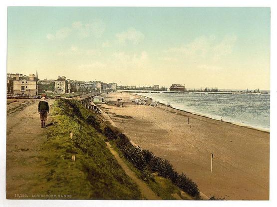 The image portrays a historical view from The Sands, situated in Lowestoft, Suffolk. This seaside locale is captured on what appears to be an old postcard or photographic print. In the foreground of this composition stands a solitary figure dressed in dark clothing with trousers and shoes that suggest early 20th-century fashion, traversing along a pathway bordered by greenery, indicative of maintained urban fringe near the beach.

The sandy stretch extends into the distance where numerous figures can be seen enjoying leisurely activities. A line of small huts or shelters is discernible at intervals on this expanse of sand, possibly serving as stalls for vendors during peak seasons. The horizon reveals a panorama with buildings that typify an early industrial-era coastal town—some are residential while others likely serve commercial functions.

The waterbody is populated by several boats and the skyline beyond stretches to include shipyards or docks indicative of Lowestoft's maritime heritage. A notable architectural feature on the far right appears as a pavilion-like structure, potentially serving social purposes for beachgoers. The skies have a warm hue which could suggest early morning or late afternoon lighting conditions.

The image is marked with text indicating it’s from "10,250 - LOWESTOFT.SANDS" and bears the watermark of "Loener," hinting at either its creator's  [...]