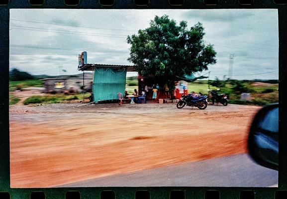A small roadside stand under a tree with people out front and a parked motorcycle. Shot from a moving car so there is motion blur.