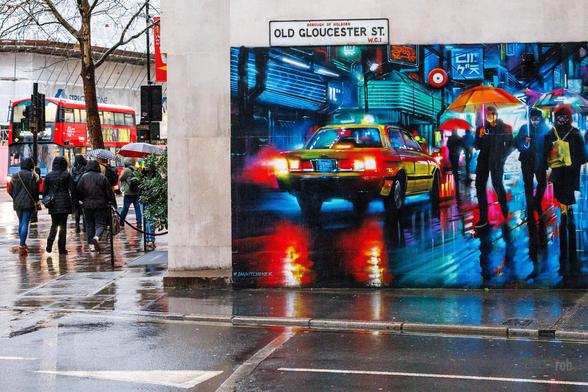 A street scene featuring a mural of yellow taxi in a vibrant rainy urban setting. Pedestrians with umbrellas walk on a wet sidewalk in the mural and on the street beside it. A red double-decker bus is visible in the background.