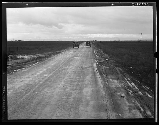 The photograph depicts a black and white scene of an unpaved rural roadway. The road stretches into the distance, with two vehicles visible on it; one appears to be in motion towards the viewer while another is further down the path ahead. Surrounding this dirt road are expansive fields that appear barren or harvested, indicating they might have been recently farmed for potatoes as suggested by a caption "Potato Field," although no actual potato plants can be seen. The sky overhead looks overcast with patches of clouds suggesting an early morning time frame. There is sparse vegetation on either side and one lone telephone pole to the right indicates some level of development or maintenance in this area. Notably, there's visible text at both top corners which seems like a recordation code for archival purposes ("3-pr821"). The overall atmosphere conveyed by the image invokes solitude typical of rural America during earlier periods, possibly mid-20th century judging from vehicle design and road condition.