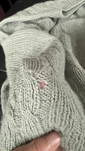 Armel eines handgestrickten Pullovers mit Zopfmuster aus salbeigrünem Alpaka-Garn. Neben einem Zopf ist ein kleines Loch in den linken Maschen.