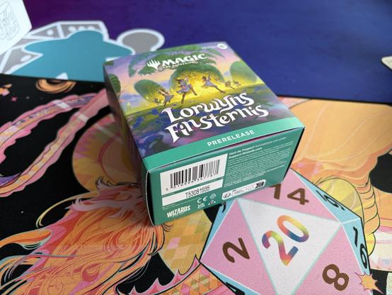 A Magic: The Gathering Lorwyn: Eclipsed prerelease box is placed on a colorful gaming mat featuring vibrant art. A twenty-sided die is also visible, indicating a tabletop gaming environment.
