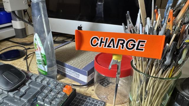 Workspace detail of a Mazda 767B model build project showing the assembled car's 'CHARGE' livery banner, surrounded by hobby tools, keyboard, and craft supplies at a maker's desk