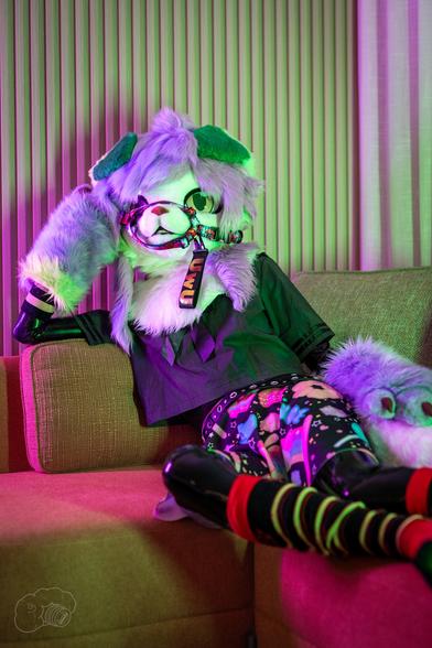 Photo by Tato Snep at Aurawra 2025.

Treble in their partial fursuit and skirt and retroreflective crop-top plus Angy paws arm warmers and leg warmers. On top of their latex catsuit.
Leaning back into the couch as they looked off in the distance.