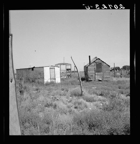 The image is a black and white photograph depicting rural, dilapidated structures. There are three main buildings visible: two appear to be old cabins or sheds made of wood with weathered exteriors, while the third has an unfinished look, perhaps indicating it's in disrepair. Surrounding these dwellings is tall grass indicative of dry conditions, possibly a field that could serve as grazing land for livestock or left uncultivated due to economic hardship.

The setting appears remote and sparsely populated with no visible signs of activity apart from the structures themselves. The lack of modern amenities like fences suggests an informal settlement without strict property boundaries.

A notable feature in this photograph is handwritten text at the top which reads "5-24ros," potentially a reference to a date or location code related to the image's creation or context within historical records, such as those from Oregon’s Klamath County. The presence of shack for potato pickers could indicate agricultural activity nearby.

The inclusion in an archive by Loener.nl suggests this photo is part of a larger collection possibly documenting living conditions and migration patterns during a specific period. Notably, the mention of a young woman from South Dakota residing in a tent house indicates that these structures are temporary dwellings for itinerant laborers or individuals dis [...]