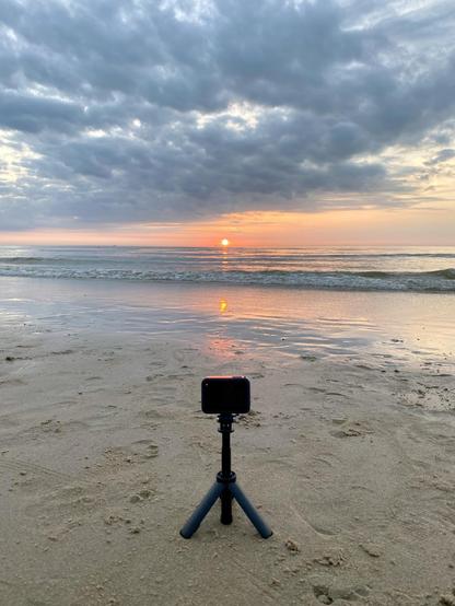 Sunrise over a calm sea, with the sun reflecting on the water. A smartphone on a tripod is set up on the sandy beach in the foreground, capturing the scene. Clouds hover above, tinged by the morning light.