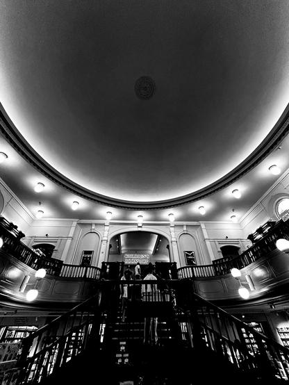 A monotone photograph of an interior with a bifurcated staircase and a domed ceiling. At the top of the stairs, through a wide arch, a sign reads "children's bookshop." On the ground floor, bookshelves can be seen through the bannisters. The building was formerly Broad Street Independent Chapel. Waterstones bookshop, Broad Street, Reading, Berkshire, UK. Photo by Gearóid Burke.