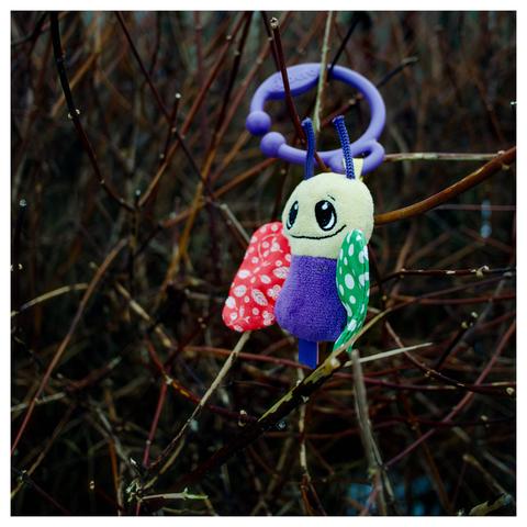 En fyrkantig bild på en leksak i plysch som hänger i en häck. Leksaken ser ut som en flerfärgat bi med en plastring.