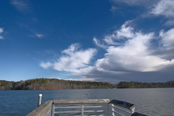 A lakeview from a deck looking at bright white clouds in a blue sky; lake water and woods on the shore