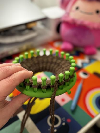 Pieni vihreä pyöreä kehikko kädessä. Kehikossa on tummanruskeaa villalankaa neulottuna, neulos on vasta alussa. Taustalla näkyy vaaleanpunainen pehmolelu. Toisen kämmekkään parin työstäminen aloitettu.