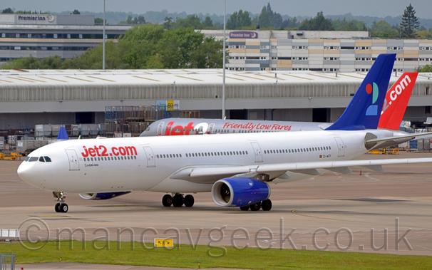 Side view of a twin engined jet  airliner taxiing from right to left along a grey concrete taxiway.
The plane is mostly white, with red "Jet2.com" titles on the upper forward fuselage, and much smaller black text "Airbus A330-300" under the forward passenger windows.
The tail is a deep blue, with a heavily stylised number "2" in red, light blue, and aquamarine.
The engine pods under the wings are the same deep blue.
In the background, another jet airliner, this one grey with red and white "Jet2" titles and red text "Friendly low fares" on the upper fuselage sits in the middle of the frame, in front of a wide, grey building that stretches the width of the frame.
A pair of larger grey buildings are in the distance, both with "Premier Inn" signs near the top.
Grey sky fills what little space is left at the top.