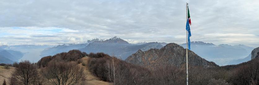 L'immagine mostra un panorama montano mozzafiato, che si estende come previsto dal Rifugio SEV (Società Escursionisti Valmadreresi) situato sull'Alpe di Pianezzo, a 1.276 metri di quota sui Corni di Canzo. 
Il primo piano è dominato da colline boscose con vegetazione spoglia, tipica della stagione invernale o autunnale. Sulla destra, un'alta asta della bandiera sventola una bandiera blu con un simbolo bianco e rosso, e la bandiera italiana in cima.
Oltre le colline, si apre un'ampia vista sul Lago di Como e sulle montagne circostanti. Le montagne, tra cui le Grigne e il Resegone, presentano cime innevate e sono avvolte da una leggera foschia o nuvole basse.
La città di Lecco è visibile sulla destra, situata sulle rive del lago, ai piedi delle montagne.
Il cielo è parzialmente nuvoloso, con chiazze di azzurro che lasciano filtrare la luce