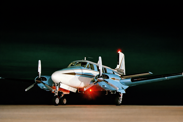 This image shows a twin-engine piston aircraft parked on a runway or ramp at night. The airplane is photographed from a front-left three-quarter angle, emphasizing its rounded nose, cockpit windows, and two spinning propellers, which appear slightly blurred. The aircraft is painted light blue and white with thin accent stripes along the fuselage. Its landing gear is extended, and red navigation and beacon lights glow beneath the fuselage and on the tail, casting small reflections on the pavement. The background is completely dark, with no visible buildings or horizon, isolating the aircraft against the night sky and drawing attention to its form and lighting.