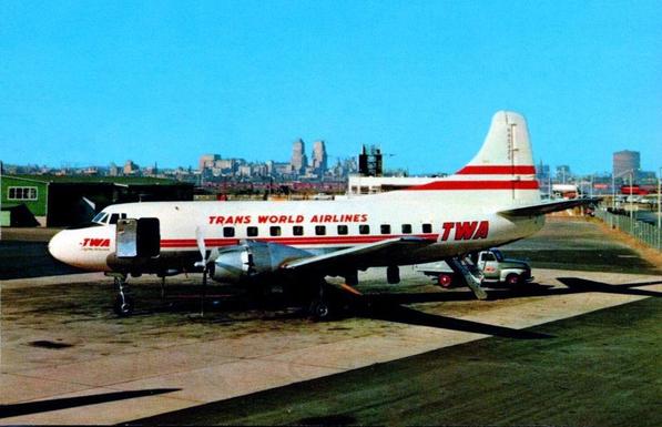 This is a color photograph of a Trans World Airlines (TWA) Martin 4-0-4 propeller aircraft parked on the airport ramp in daylight. The airplane is shown in full side view, facing left. It is painted white with bold red stripes and large red lettering reading “TRANS WORLD AIRLINES” along the fuselage, with “TWA” on the tail and near the nose.

The aircraft has two propeller engines mounted on low wings, a rounded nose, and a tall vertical tail. A small set of mobile stairs is positioned at the rear passenger door. The landing gear is extended, and the plane rests on the concrete apron.

In the background, airport buildings and hangars sit low along the horizon, with a distant city skyline visible under a clear blue sky. The scene feels calm and orderly, typical of a mid-20th-century commercial airport setting.