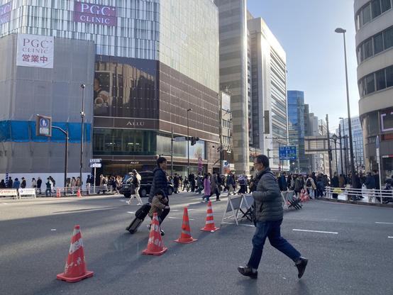 A busy urban street scene featuring pedestrians crossing the road. Various office buildings and shops are visible, with construction barriers and traffic signs present. Some individuals are carrying luggage, and many people are wearing winter clothing.
