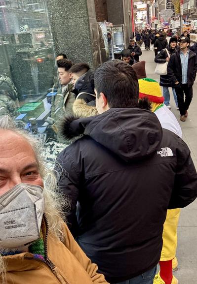 A selfie of us on line to get in the gold & silver store. The display case is partly seen, and people ahead of me in line. One man’s color-themed outfit is partially seen, red socks/ yellow sneakers and a yellow/ green/ red hat.