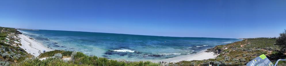 Panorama view of the Iluka beach and surrounds.  To the left is an expansive boat harbour reaching out to the bright blue and green ocean. To the right are wide patches of native bushland leading to several beaches.  A lone cyclepath winds through the dunes like a snake.  The sky is a majestic blue free of clouds.