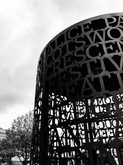 A large, cylindrical metal installation consisting of 18 lines of text in circles, stacked on top of each other. It features names of local streets, neighbourhoods, and cultural institutions.  Southgate, Bath, UK. Photo by Gearóid Burke.