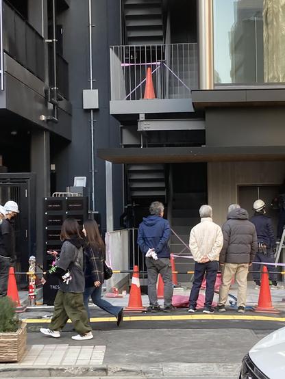 The image shows a street scene with several people observing a construction area. There are traffic cones, construction materials, and a set of stairs in the background. A group of three men stands watching, while two women walk past in the foreground.