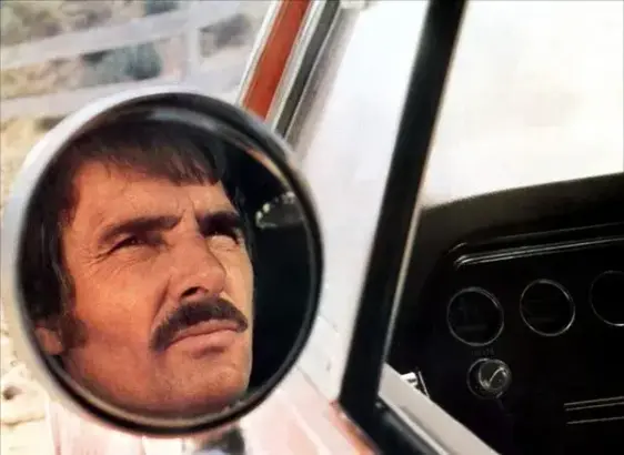A still from the film Duel. A moustachioed white man's face can be seen in the round wing mirror of a car.