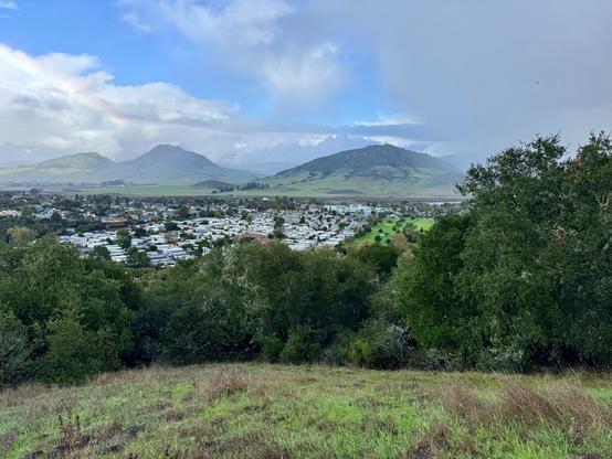 While standing on one hill, the view is looking across a slight valley of houses to the other side wit three mountain peaks and soft clouds overhead; some spots are raining, and a very soft rainbow is seen in the upper left. Green grass and trees/large bushes are seen in the foreground. Sterling trail in the Irish Hills, San Luis Obispo, California