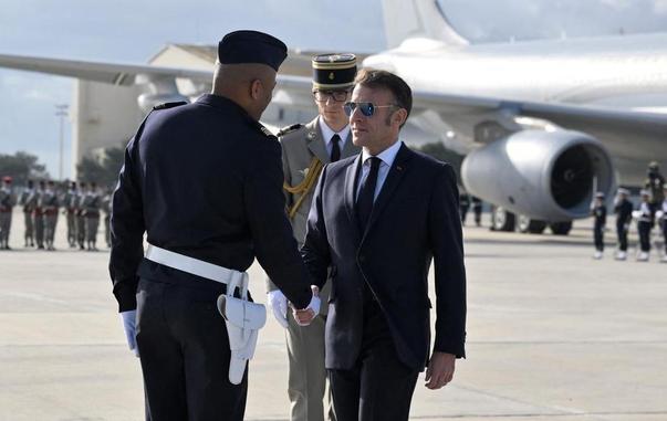 Photo d'Emmanuel Macron qui serre la main d'un officier de l'armée de l'air. En fonds on voit un gros porteur. Macron a des lunettes aviator.