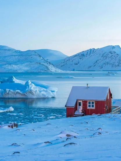 A serene winter scene featuring a red cabin by icy waters, surrounded by snow-covered mountains and floating icebergs under a blue sky.